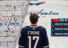 Hockey player Dylan Strome seen from behind, wearing his #17 Washington Capitals jersey while standing at center ice and looking at the team logo. A graphic overlay on the right displays his projected 2025-26 stats: 32 goals, 54 assists, and 86 points.