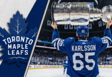 Erik Karlsson of the Toronto Maple Leafs, viewed from behind wearing his #65 jersey, hoists the Stanley Cup above his head at center ice in a packed Scotiabank Arena.