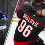 Jack Roslovic in his Carolina Hurricanes uniform, looking on during an NHL game. The background shows the ice and stands and the Canucks logo to the left