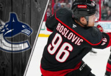 Jack Roslovic in his Carolina Hurricanes uniform, looking on during an NHL game. The background shows the ice and stands and the Canucks logo to the left
