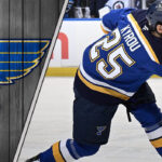 St. Louis Blues forward Jordan Kyrou takes a slapshot during an NHL game, with the Blues logo visible in the background.