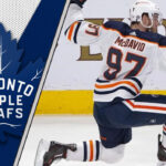 Connor McDavid celebrates a goal during a game, with the Toronto Maple Leafs logo in the background, fueling NHL rumors of a potential trade.