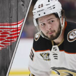 An image of Mason McTavish in his Anaheim Ducks jersey, looking focused during a game, with the Detroit Red Wings logo in the background.