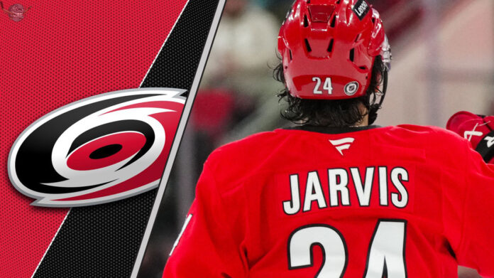 Seth Jarvis, forward for the Carolina Hurricanes, skates with the puck during a game. He is wearing a red Hurricanes jersey with the number 24. The background is blurred, showing the ice and boards of a hockey arena. He is poised for a breakout season according to fantasy hockey projections.