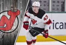 New Jersey Devils defenseman Luke Hughes skates with the puck during an NHL game, with the Devils logo in the background.