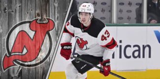 New Jersey Devils defenseman Luke Hughes skates with the puck during an NHL game, with the Devils logo in the background.