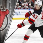 New Jersey Devils defenseman Luke Hughes skates with the puck during an NHL game, with the Devils' logo visible in the background.