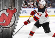 New Jersey Devils defenseman Luke Hughes skates with the puck during an NHL game, with the Devils' logo visible in the background.