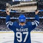 Image of hockey player Connor McDavid celebrating at center ice, wearing a Toronto Maple Leafs jersey with his name and number 97. He is smiling and hoisting the Stanley Cup above his head as blue and white confetti falls in a packed arena.