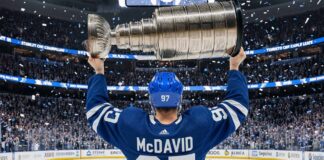 Image of hockey player Connor McDavid celebrating at center ice, wearing a Toronto Maple Leafs jersey with his name and number 97. He is smiling and hoisting the Stanley Cup above his head as blue and white confetti falls in a packed arena.