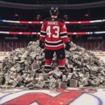 Luke Hughes of the New Jersey Devils Stands in front of a giant pile of money on the ice at his home arena.