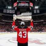 A wide-angle photograph from behind shows a New Jersey Devils hockey player wearing jersey number 43 with the name "HUGHES" across the back, holding the silver Stanley Cup trophy overhead with both hands at center ice of a packed Prudential Center, while the Jumbotron above reads "DEVILS STANLEY CUP CHAMPIONS."