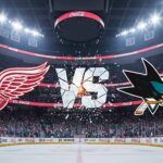Can the Red Wings Bounce Back in San Jose? How to Watch, Key Injuries & Game Predictions A stylized image depicting the Detroit Red Wings logo and the San Jose Sharks logo colliding above center ice in a packed hockey arena. The word 'VS' shatters between them, illuminated by spotlights in the background, which is the San Jose Sharks' home arena.