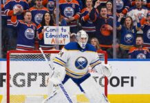 Buffalo Sabres goaltender Alex Lyon (#34), wearing his white away jersey, stands in a ready position in front of his net. Behind the protective glass, the stands are filled mostly with cheering Edmonton Oilers fans in blue and orange jerseys.