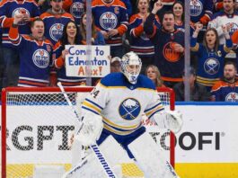 Buffalo Sabres goaltender Alex Lyon (#34), wearing his white away jersey, stands in a ready position in front of his net. Behind the protective glass, the stands are filled mostly with cheering Edmonton Oilers fans in blue and orange jerseys.