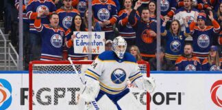 Buffalo Sabres goaltender Alex Lyon (#34), wearing his white away jersey, stands in a ready position in front of his net. Behind the protective glass, the stands are filled mostly with cheering Edmonton Oilers fans in blue and orange jerseys.
