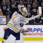 Buffalo Sabres goaltender Alex Lyon (34), wearing a white away jersey, crouches on the ice looking towards the play. Behind the glass in the background, Los Angeles Kings fans watch, with one woman holding a handwritten sign that reads, "WE WANT YOU ALEX!".