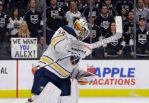 Buffalo Sabres goaltender Alex Lyon (34), wearing a white away jersey, crouches on the ice looking towards the play. Behind the glass in the background, Los Angeles Kings fans watch, with one woman holding a handwritten sign that reads, "WE WANT YOU ALEX!".