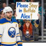 Alex Tuch Wants Out? The $10M Demand Shocking Sabres Fans Buffalo Sabres forward Alex Tuch (89) stands on the ice during an away game, looking towards the left. Directly behind him behind the rink glass, a smiling female fan with long red hair, wearing a Sabres jersey and backwards hat, holds up a large handmade white sign with blue and gold lettering that reads, "Please Stay in Buffalo Alex!".