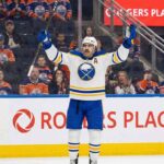 Buffalo Sabres alternate captain Alex Tuch (#89), wearing a white away jersey, raises his arms in celebration while standing on the ice at Rogers Place. Behind him, the stands are filled with seated fans wearing Edmonton Oilers jerseys.