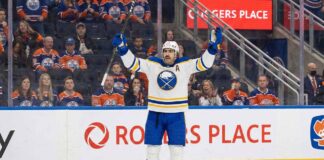 Buffalo Sabres alternate captain Alex Tuch (#89), wearing a white away jersey, raises his arms in celebration while standing on the ice at Rogers Place. Behind him, the stands are filled with seated fans wearing Edmonton Oilers jerseys.