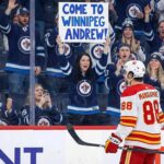Calgary Flames forward Andrew Mangiapane (88), wearing a white away jersey, stands on the ice looking towards the spectator stands behind the glass. A female fan in the front row, wearing a blue Winnipeg Jets jersey, holds up a large white sign with blue lettering that reads, "COME TO WINNIPEG ANDREW!". The surrounding crowd is mostly cheering and wearing Jets apparel.