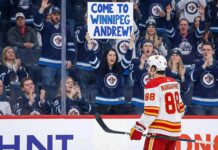 Calgary Flames forward Andrew Mangiapane (88), wearing a white away jersey, stands on the ice looking towards the spectator stands behind the glass. A female fan in the front row, wearing a blue Winnipeg Jets jersey, holds up a large white sign with blue lettering that reads, "COME TO WINNIPEG ANDREW!". The surrounding crowd is mostly cheering and wearing Jets apparel.