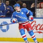 New York Rangers alternate captain Artemi Panarin (10) skating intently on the ice during a game. In the stands behind the glass, a red-haired fan holds up a white sign manually written with "WE WANT TWO FIRST-ROUND PICKS FOR PANARIN".