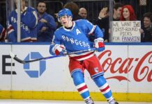 New York Rangers alternate captain Artemi Panarin (10) skating intently on the ice during a game. In the stands behind the glass, a red-haired fan holds up a white sign manually written with "WE WANT TWO FIRST-ROUND PICKS FOR PANARIN".