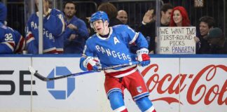 New York Rangers alternate captain Artemi Panarin (10) skating intently on the ice during a game. In the stands behind the glass, a red-haired fan holds up a white sign manually written with "WE WANT TWO FIRST-ROUND PICKS FOR PANARIN".