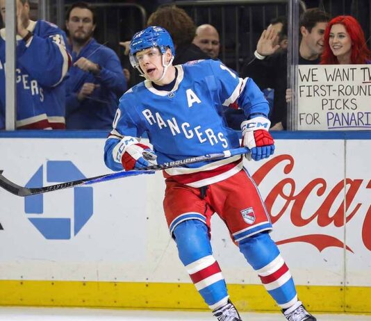New York Rangers alternate captain Artemi Panarin (10) skating intently on the ice during a game. In the stands behind the glass, a red-haired fan holds up a white sign manually written with "WE WANT TWO FIRST-ROUND PICKS FOR PANARIN".