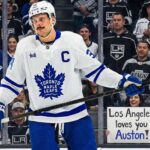 Toronto Maple Leafs captain Auston Matthews stands on the ice in his white away jersey, looking towards the stands behind the glass. The crowd features numerous Los Angeles Kings fans, and a smiling woman in the front row holds up a handwritten sign that reads, "Los Angeles loves you Auston!", amidst ongoing trade speculation.