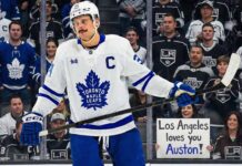 Toronto Maple Leafs captain Auston Matthews stands on the ice in his white away jersey, looking towards the stands behind the glass. The crowd features numerous Los Angeles Kings fans, and a smiling woman in the front row holds up a handwritten sign that reads, "Los Angeles loves you Auston!", amidst ongoing trade speculation.