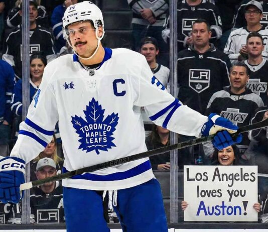 Toronto Maple Leafs captain Auston Matthews stands on the ice in his white away jersey, looking towards the stands behind the glass. The crowd features numerous Los Angeles Kings fans, and a smiling woman in the front row holds up a handwritten sign that reads, "Los Angeles loves you Auston!", amidst ongoing trade speculation.