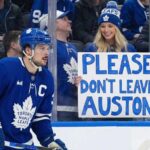 Auston Matthews, wearing his blue Toronto Maple Leafs jersey with a captain's 'C', is on the ice during a game, looking towards the stands where a smiling blonde woman holds a handmade sign that reads "PLEASE DON'T LEAVE AUSTON".