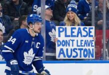 Auston Matthews, wearing his blue Toronto Maple Leafs jersey with a captain's 'C', is on the ice during a game, looking towards the stands where a smiling blonde woman holds a handmade sign that reads "PLEASE DON'T LEAVE AUSTON".