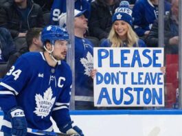 Auston Matthews, wearing his blue Toronto Maple Leafs jersey with a captain's 'C', is on the ice during a game, looking towards the stands where a smiling blonde woman holds a handmade sign that reads "PLEASE DON'T LEAVE AUSTON".
