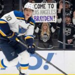 St. Louis Blues captain Brayden Schenn (#10) skates with the puck past tall rink boards featuring a Coca-Cola advertisement. Behind the glass, Los Angeles Kings fans cheer, and a woman holds up a handmade sign that reads "COME TO LOS ANGELES BRAYDEN!".