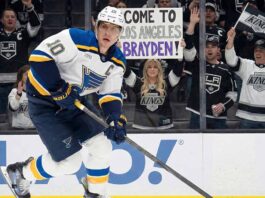 St. Louis Blues captain Brayden Schenn (#10) skates with the puck past tall rink boards featuring a Coca-Cola advertisement. Behind the glass, Los Angeles Kings fans cheer, and a woman holds up a handmade sign that reads "COME TO LOS ANGELES BRAYDEN!".