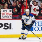 St. Louis Blues forward Brayden Schenn, wearing his white away jersey, skates with the puck during a game. In the stands behind the glass, a crowd of Ottawa Senators fans cheer, and a blonde woman in the front row holds up a white sign with red and black text that reads, "COME TO OTTAWA BRAYDEN".