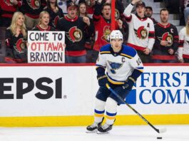 St. Louis Blues forward Brayden Schenn, wearing his white away jersey, skates with the puck during a game. In the stands behind the glass, a crowd of Ottawa Senators fans cheer, and a blonde woman in the front row holds up a white sign with red and black text that reads, "COME TO OTTAWA BRAYDEN".
