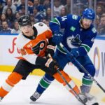 Philadelphia Flyers' Trevor Zegras and Vancouver Canucks' Kiefer Sherwood battle for the puck along the boards during an NHL game.