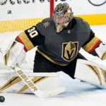 Carter Hart, in a grey home jersey and custom gold-and-black helmet is down in a butterfly position on the ice, making a stick save on a puck immediately in front of the goal crease.