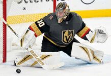 Carter Hart, in a grey home jersey and custom gold-and-black helmet is down in a butterfly position on the ice, making a stick save on a puck immediately in front of the goal crease.