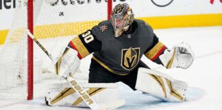 Carter Hart, in a grey home jersey and custom gold-and-black helmet is down in a butterfly position on the ice, making a stick save on a puck immediately in front of the goal crease.