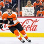 Why The Philadelphia Flyers Are Scrambling to Lock Down Christian Dvorak Before July Philadelphia Flyers forward Christian Dvorak (wearing number 22) skating with the puck during an NHL game. In the background stands, a female fan holds up a handwritten sign that reads, "Please stay in Philly Christian!" amid other fans in orange jerseys.
