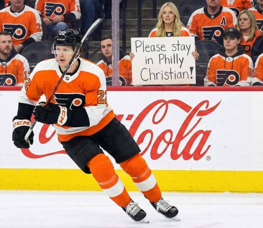 Why The Philadelphia Flyers Are Scrambling to Lock Down Christian Dvorak Before July Philadelphia Flyers forward Christian Dvorak (wearing number 22) skating with the puck during an NHL game. In the background stands, a female fan holds up a handwritten sign that reads, "Please stay in Philly Christian!" amid other fans in orange jerseys.