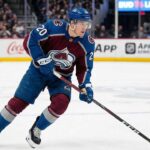 Colorado Avalanche center Ross Colton, wearing number 20 in a burgundy and blue home jersey and a blue helmet, skates across the ice with his stick down during an NHL game.