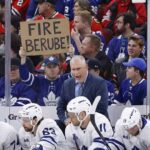 Toronto Maple Leafs head coach Craig Berube yells angrily from behind the players' bench during a game. Directly behind him in the stands, a fan holds up a handwritten cardboard sign that explicitly reads "FIRE BERUBE!".