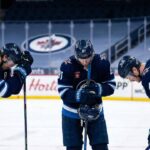Winnipeg Jets players Mark Scheifele (#55), Kyle Connor (#81), and Josh Morrissey (#44) stand dejected on the ice with their heads bowed in frustration inside an empty arena.